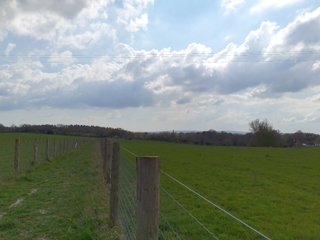 View South along the route, showing a field of grass and trees in the distance.  The sky is a little cloudy.